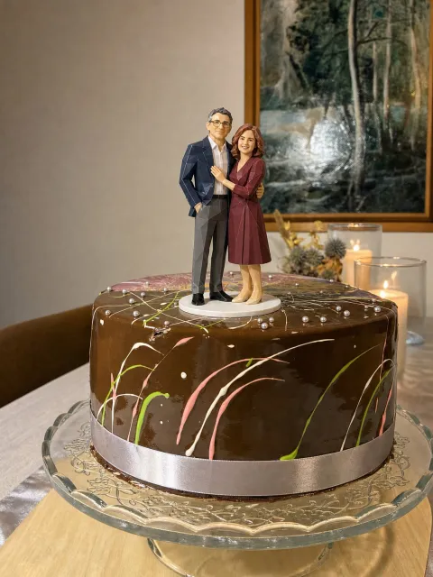 Faceted full-color figure of a couple in their fifties, he in a navy jacket and glasses, she in a burgundy dress, on a chocolate cake with a silver ribbon.