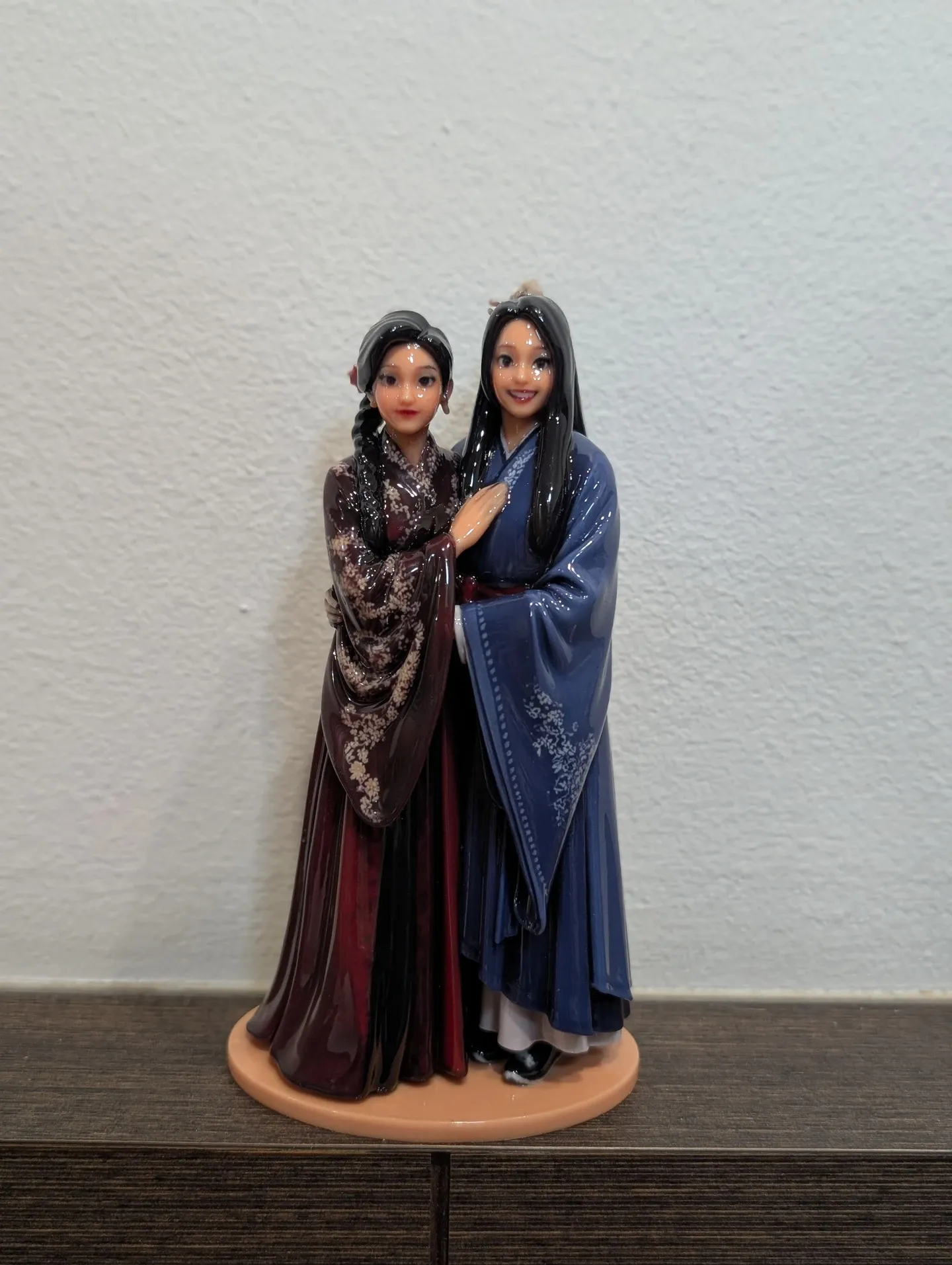 Glossy printed figure of the same two people in red and blue robes, front view, on an oval base on a dark wood sideboard