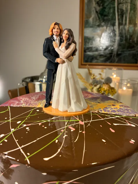 Glossy printed figure of a groom in a dark suit and a bride in a white gown, hugging, on an orange base on top of a chocolate cake with colored drizzle