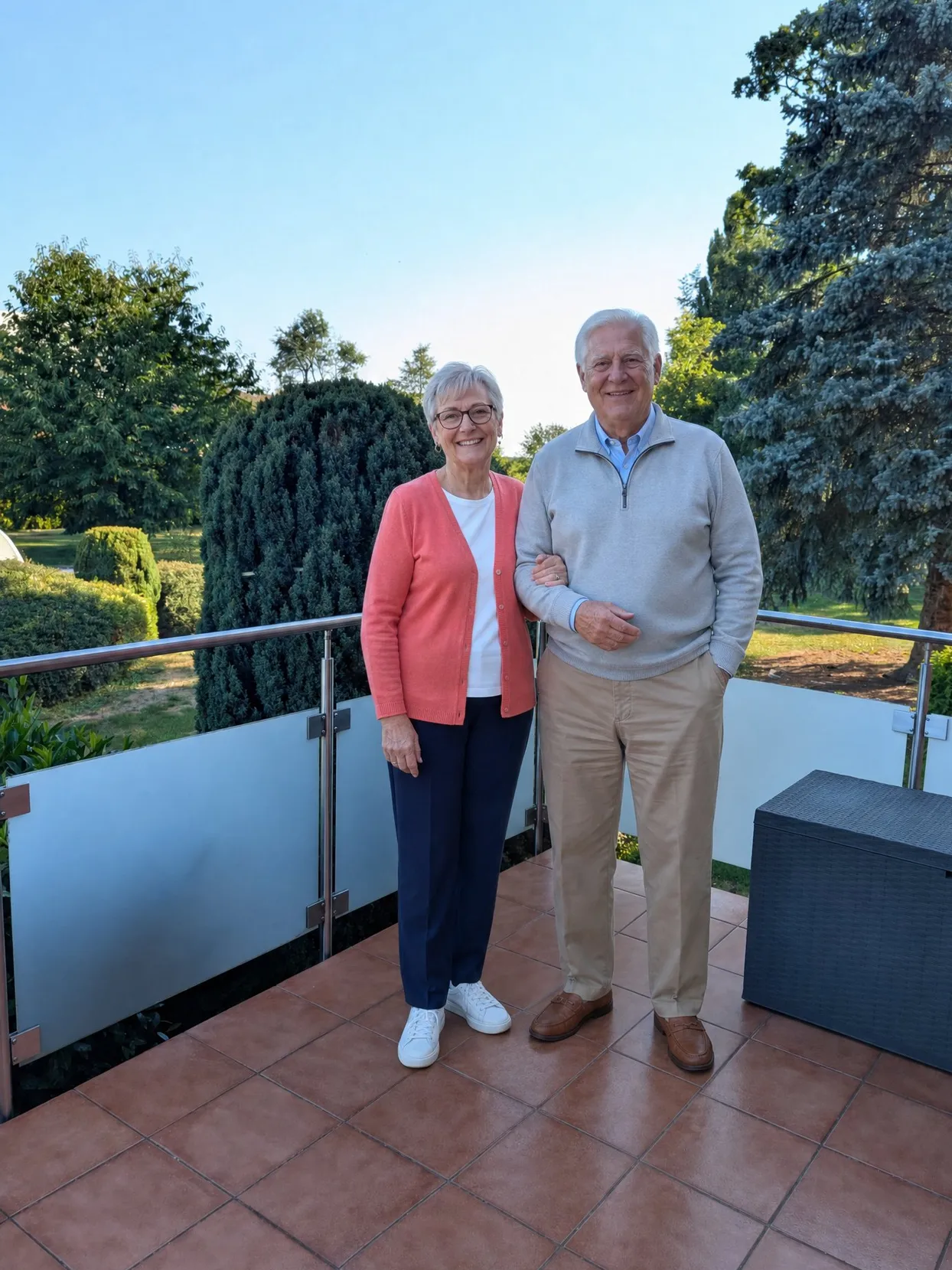 Older couple standing side by side on a sunny terrace, both seen head to toe.