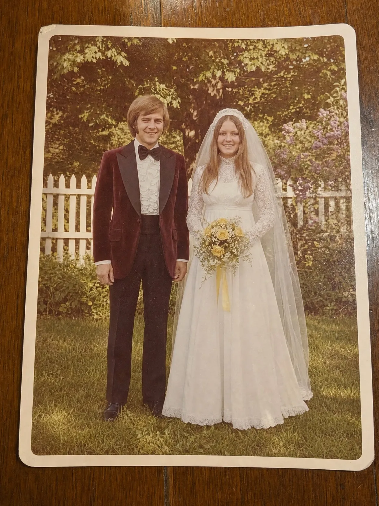 Faded wedding photo from the 1970s: groom in a burgundy velvet tuxedo, bride in a high-neck lace gown and veil with yellow roses, on a lawn by a picket fence.