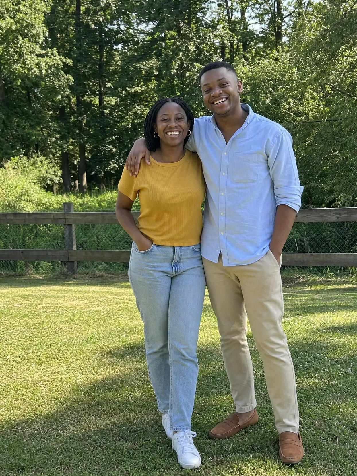 A young couple on a sunny lawn by a wooden fence, she in a mustard top and jeans, he in a light blue shirt and beige chinos, his arm around her shoulders.
