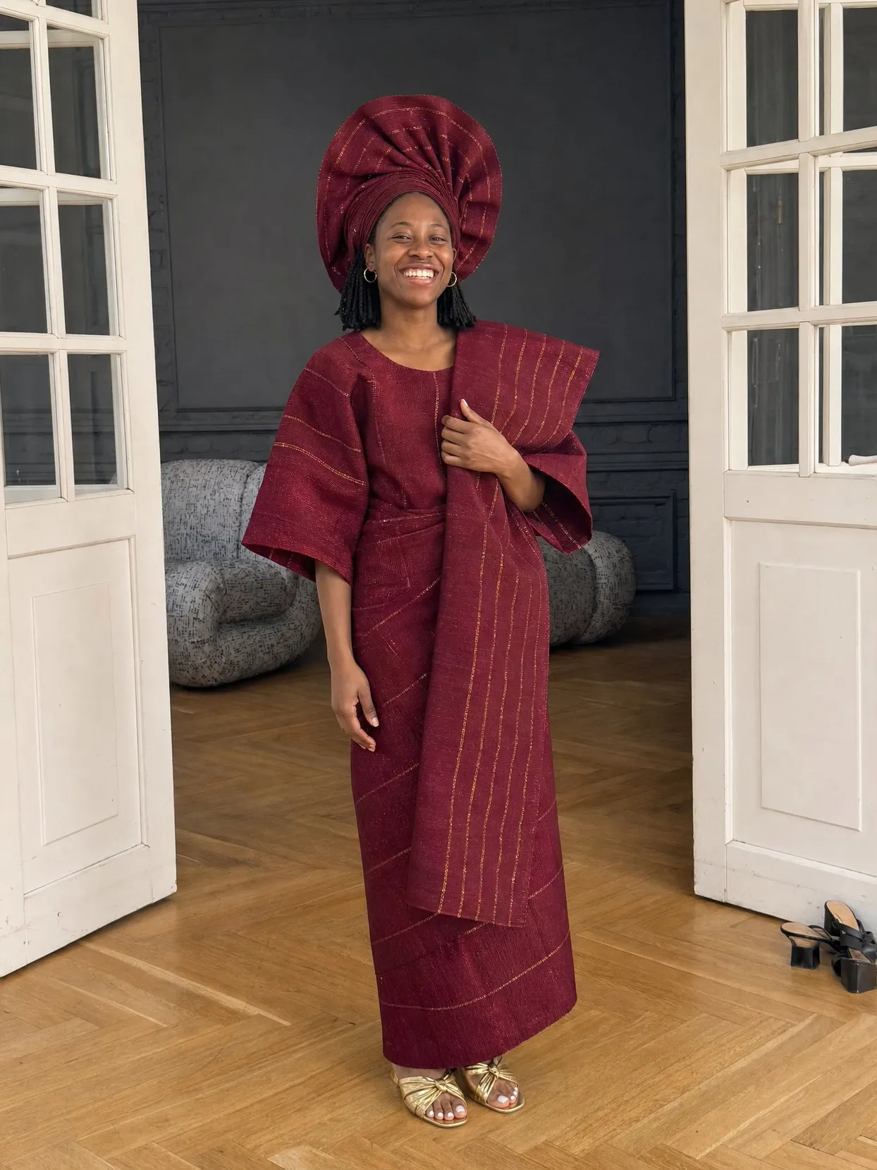 The same woman at home between open white doors, in a wine-red aso oke buba, iro and ipele with a tall matching gele.