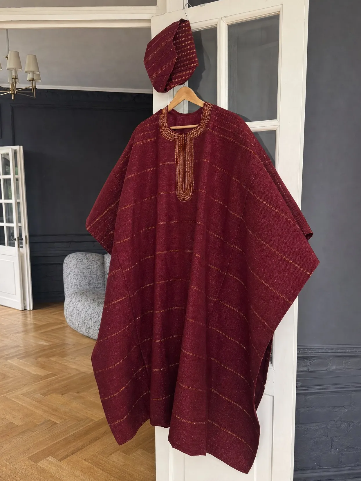 A wine-red aso oke agbada with gold embroidery at the neck and a matching fila cap, on a hanger on an open white door.