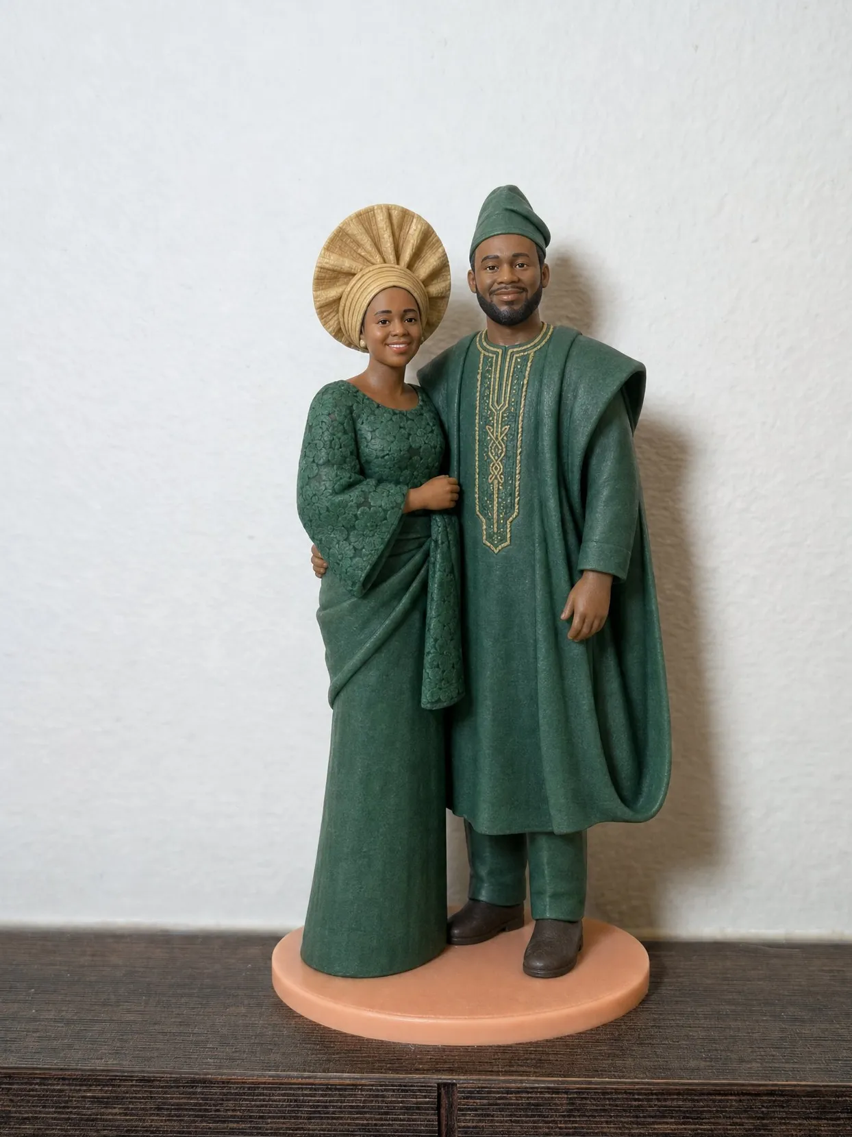 The same couple as a topper on a peach base on a wooden shelf, now in emerald green. She wears green lace with a gold gele. He wears a green agbada and fila.