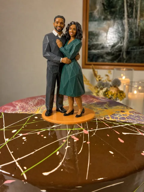 Glossy faceted figure of a couple embracing, he in a gray suit, she in a teal wrap dress, on a chocolate glaze cake.
