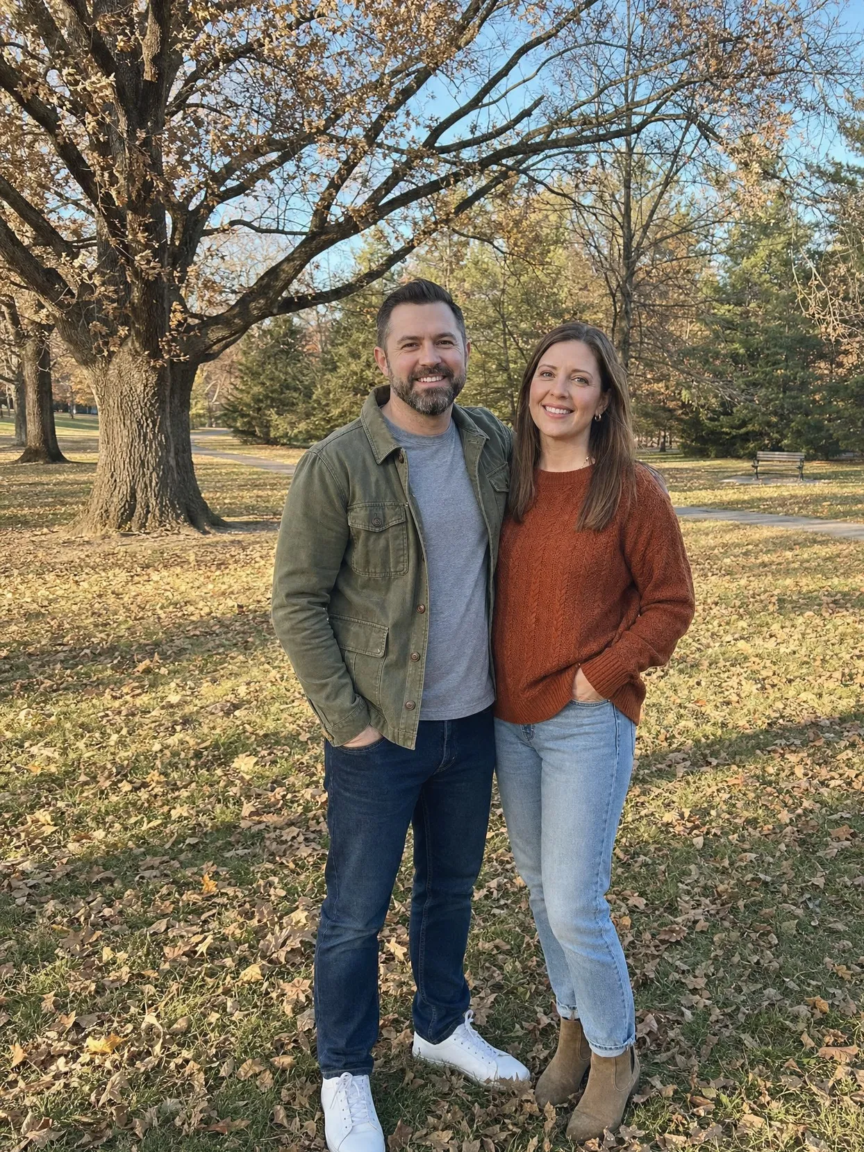 A man in an olive jacket, gray T-shirt and dark jeans and a woman in a rust sweater and light jeans stand side by side on a leafy park lawn.