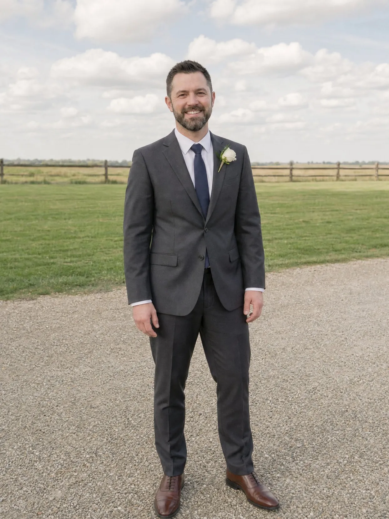 The same man in a charcoal suit, white shirt, navy tie and a white rose boutonniere, standing on a gravel drive.