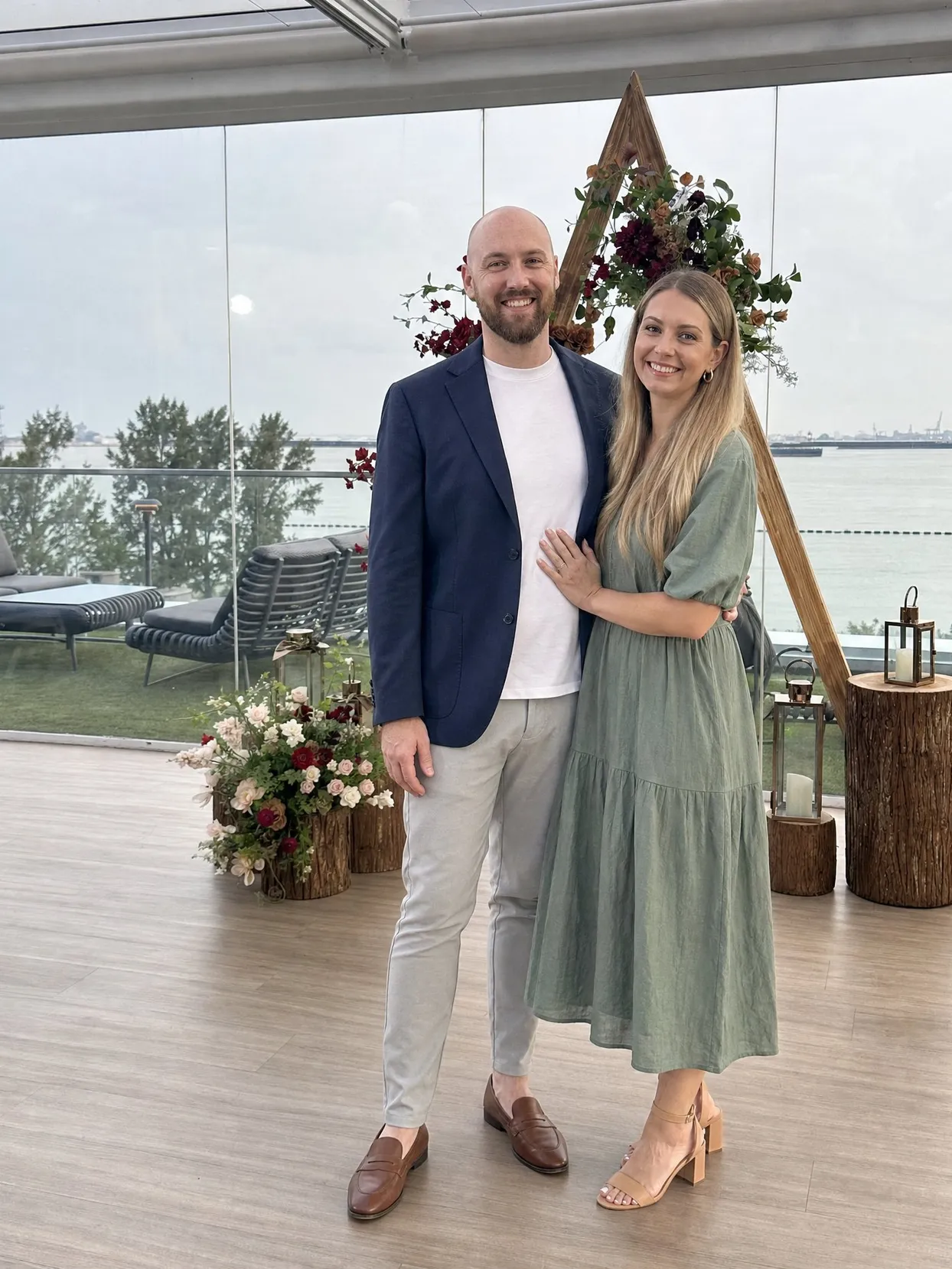 A couple on a wooden deck in a glass-walled venue by the water: he has a shaved head and a short brown beard, a navy blazer, white T-shirt and light grey chinos; she has long blonde hair and a sage-green midi dress