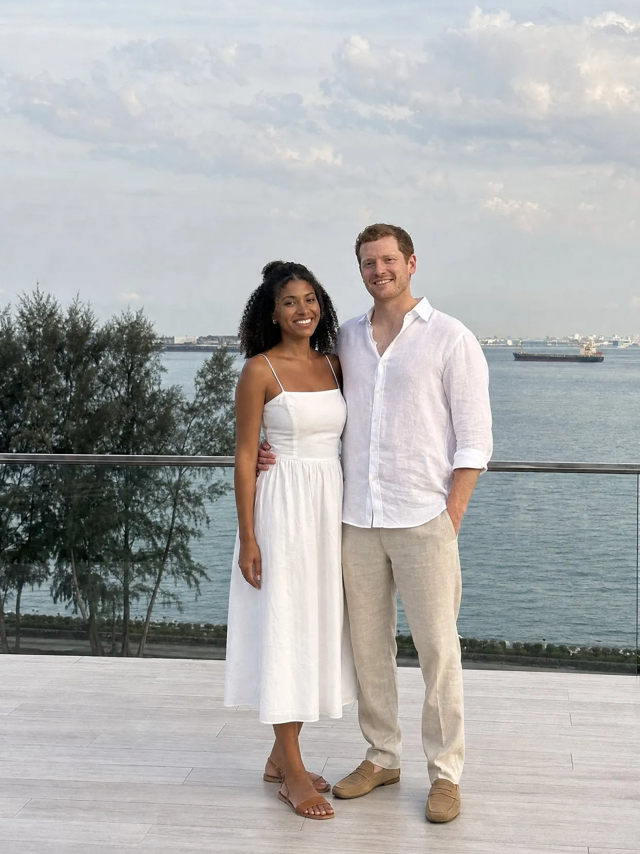 A couple on a deck by the sea, she in a white summer dress and sandals, he in a white linen shirt and light linen trousers