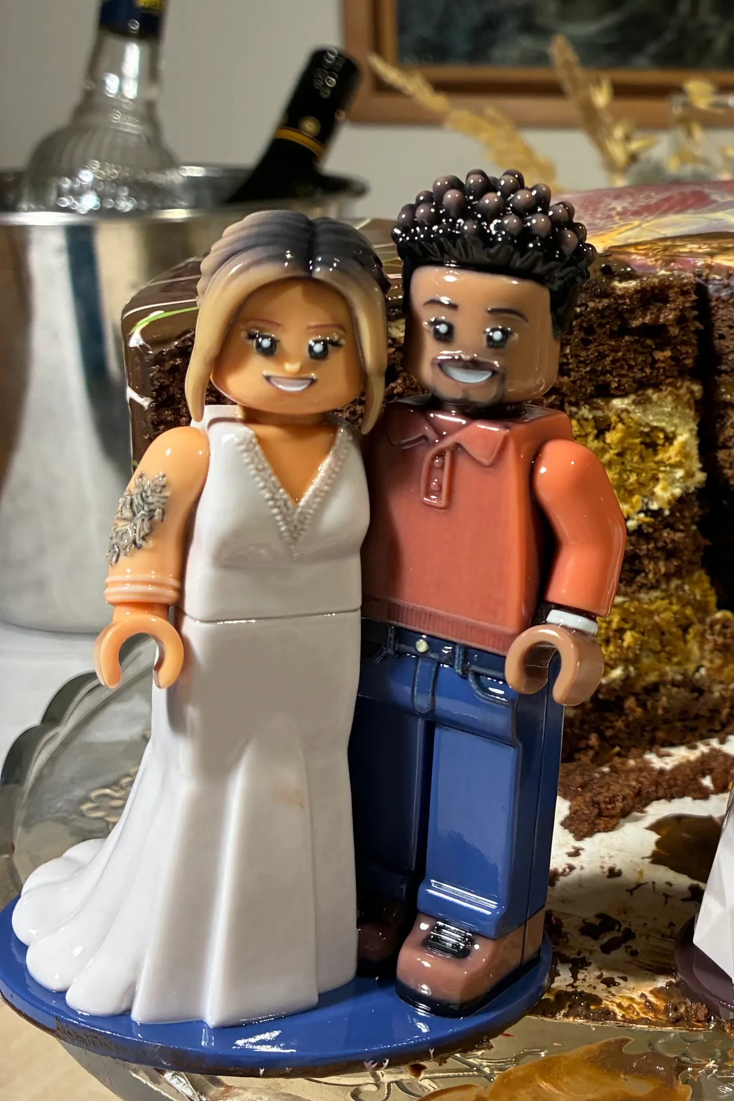 Lesklá figurka na dort ve stylu Brick Figures na nakrojeném čokoládovém dortu: nevěsta v bílých šatech s tetováním na paži, ženich v oranžové košili a džínách.
