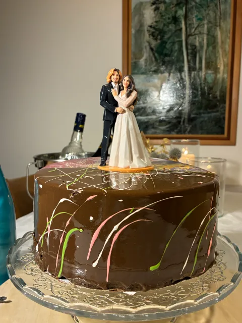 Figure of a groom in a black suit and a bride in a white gown on a chocolate cake with splatter glaze, on a glass stand, with candles and dried flowers behind