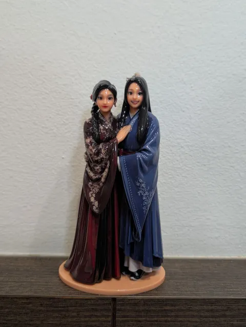 Glossy printed couple in long red and blue robes standing on a round base on a dark wood sideboard against a white wall