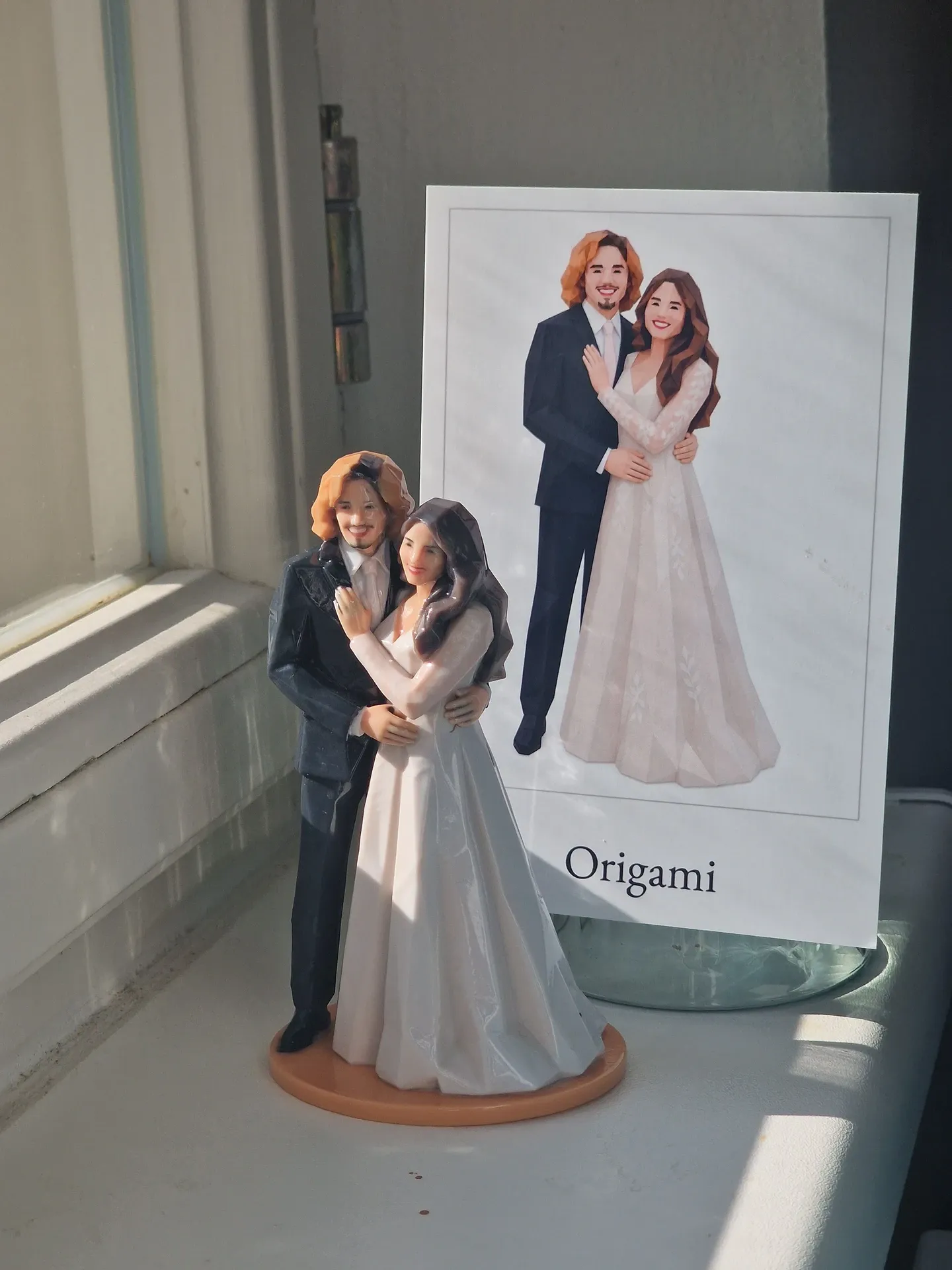 Printed bride and groom figure on an orange base on a sunny window sill, a card with a drawing of the couple behind it