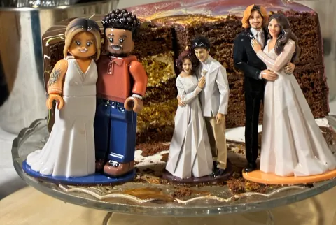 Three printed couple figures on round bases, standing on a glass cake stand in front of a cut chocolate layer cake