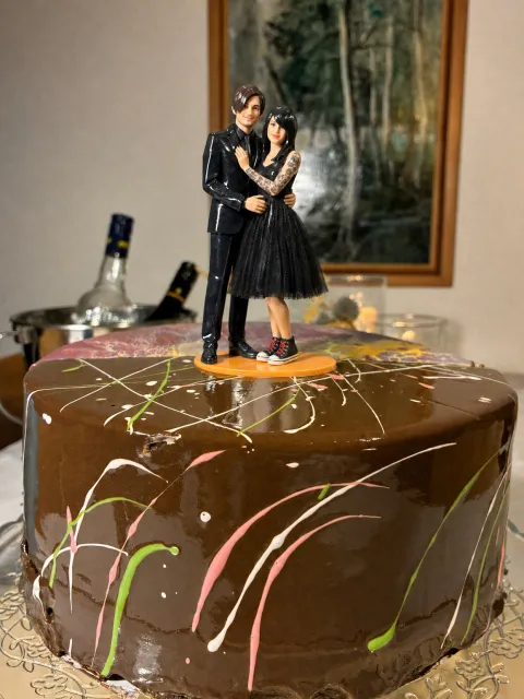 Glossy na topper ng magkayakap na couple sa orange na base sa chocolate cake na may makulay na drizzle. Ang babae ay may tuwid na itim na buhok na may mahabang side fringe, tattoo sleeve, itim na tulle dress at itim na high-tops. Ang lalaki ay may madilim na side fringe, black suit, itim na shirt at skinny na itim na tie.