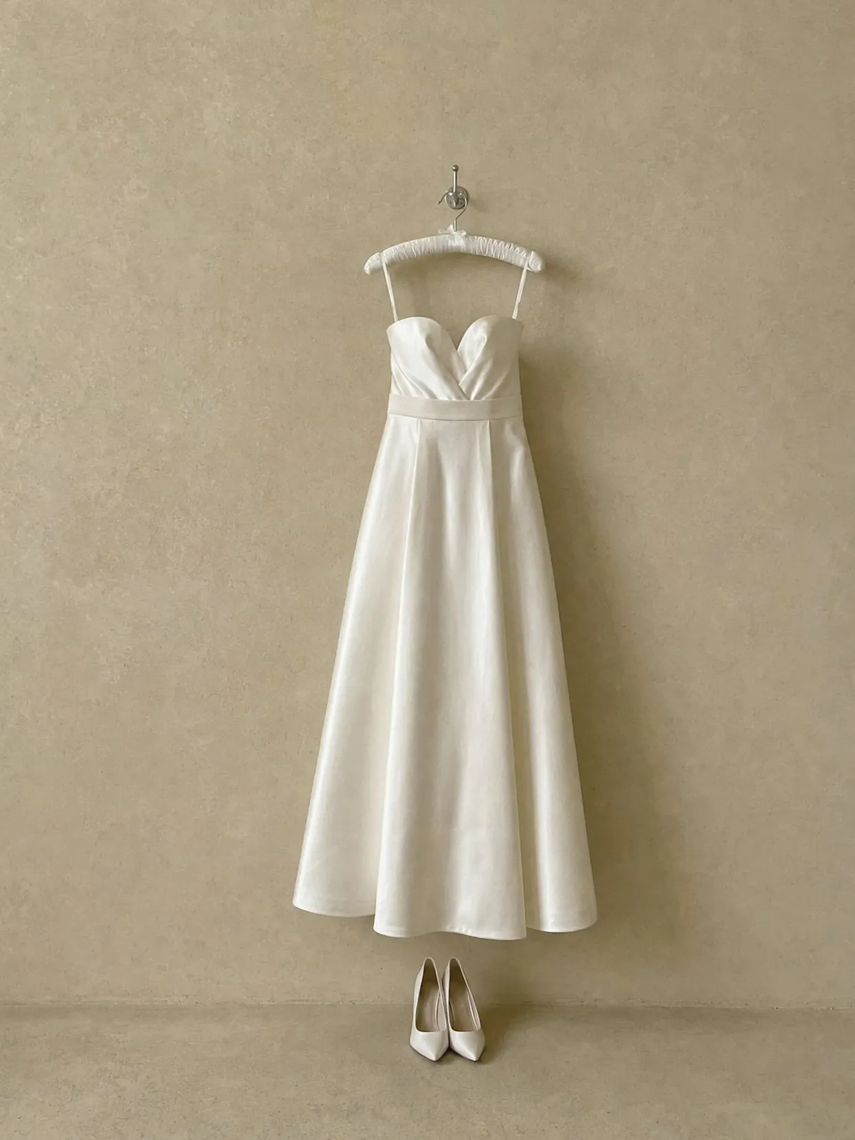 An ivory satin A-line wedding gown with a sweetheart neckline on a padded hanger on a beige wall, ivory heels on the floor below.