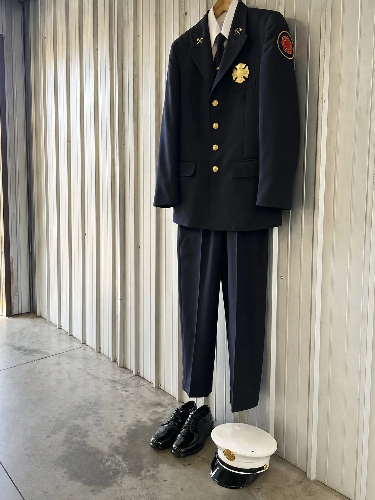 A navy firefighter dress uniform with gold buttons and a red shoulder patch, on a hanger on a metal wall. Black shoes and a white dress cap on the floor below.