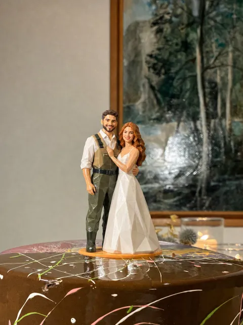 An Origami topper on a chocolate drizzle cake. The groom wears chest waders with straps over a white shirt. The bride wears a white gown.