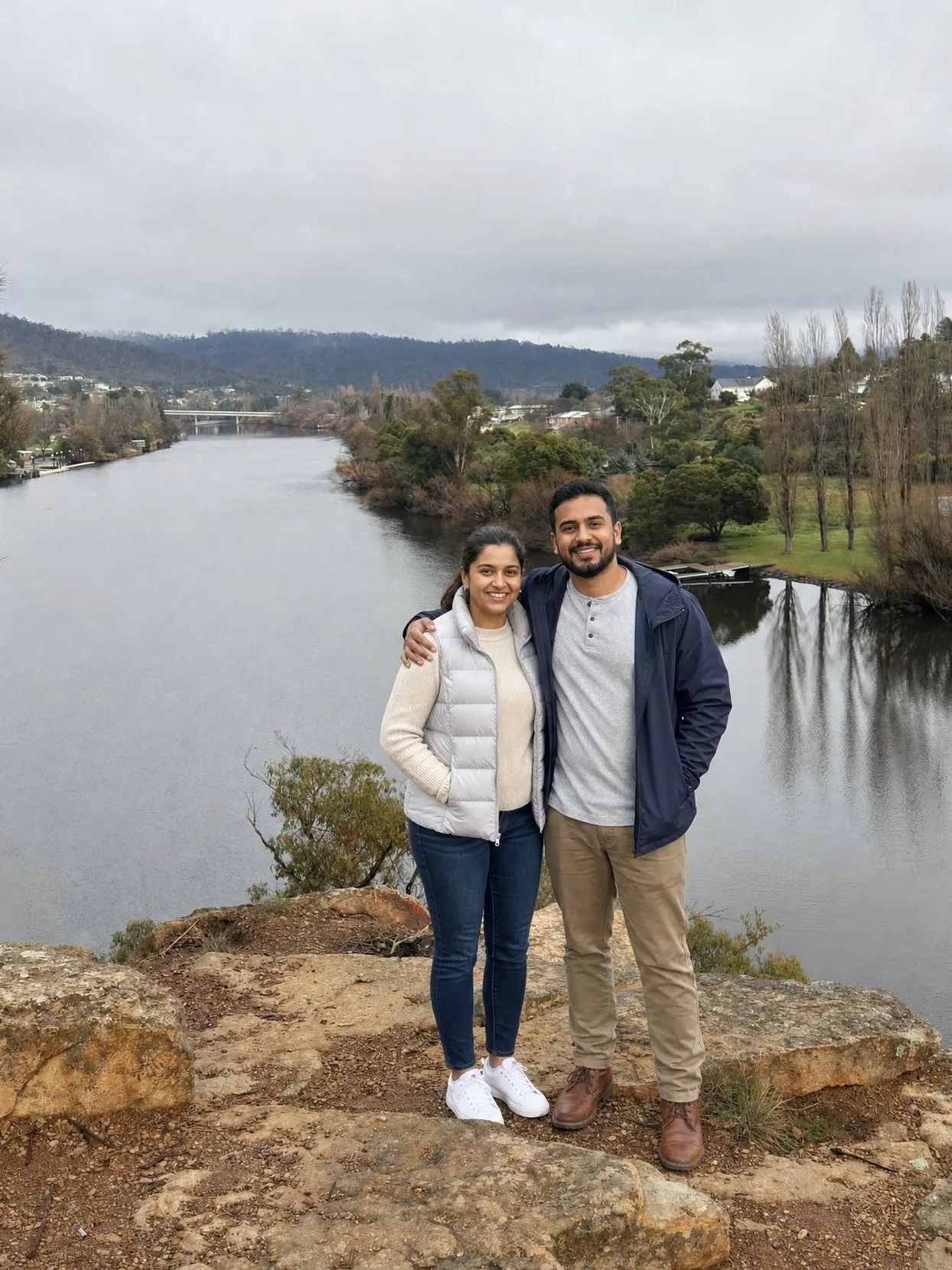 A couple on a rocky lookout above a river, she in a cream sweater and gray vest, he in a navy jacket and khakis.