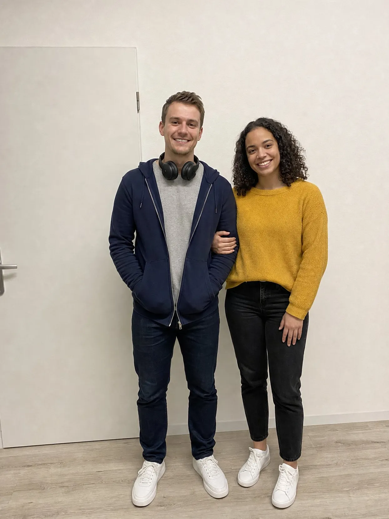 A couple standing against a white wall, he in a navy hoodie with headphones around his neck, she in a yellow sweater.