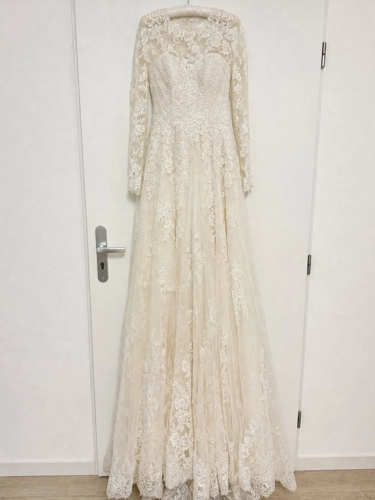 An ivory lace wedding gown with long lace sleeves on a hanger on a white door.