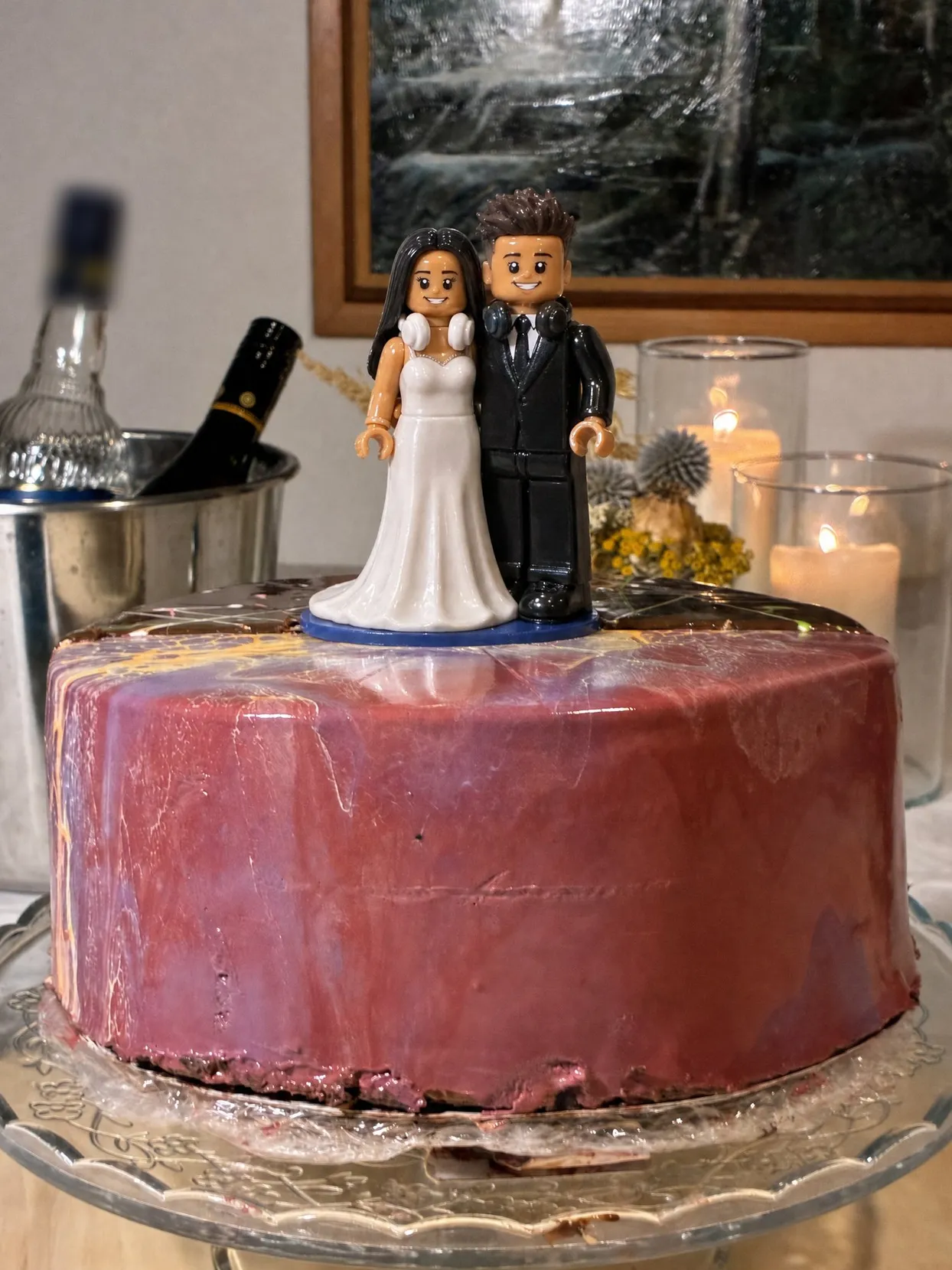 A couple topper in a white gown and a black suit, both with headphones around their necks, on a glazed cake on a glass stand.