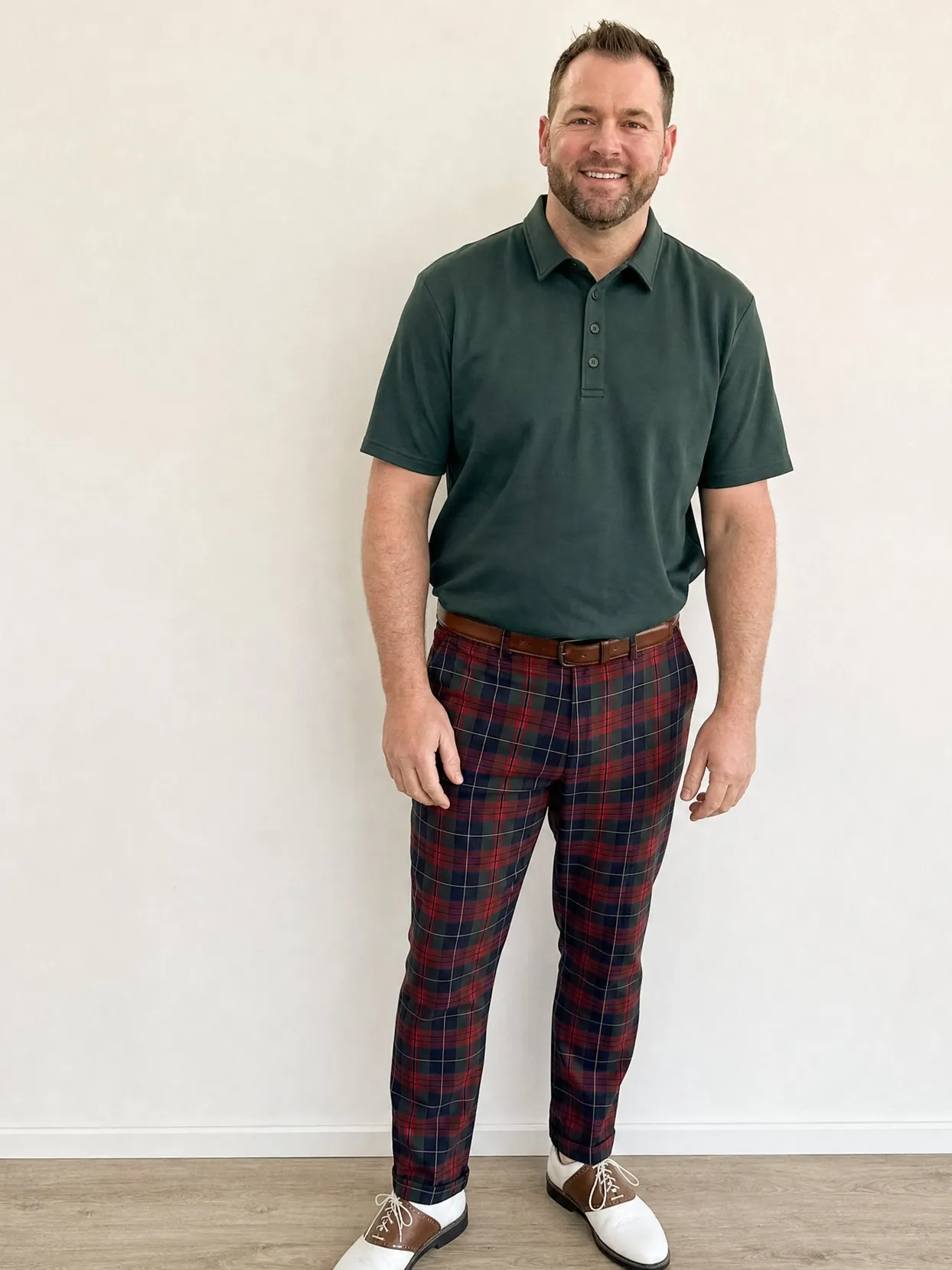 The man from the couple photo in the same polo, trousers and shoes, against a plain wall.