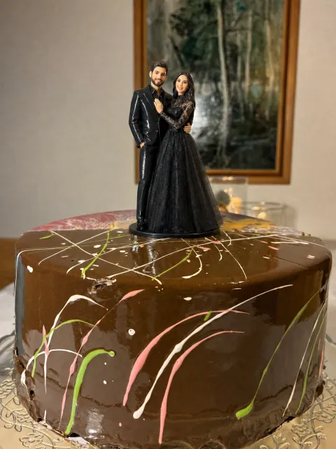 Glossy topper of a couple on a black base on a chocolate drip cake. She wears a black corset gown with a high lace neck and long black lace sleeves. He wears a black suit and a black shirt.