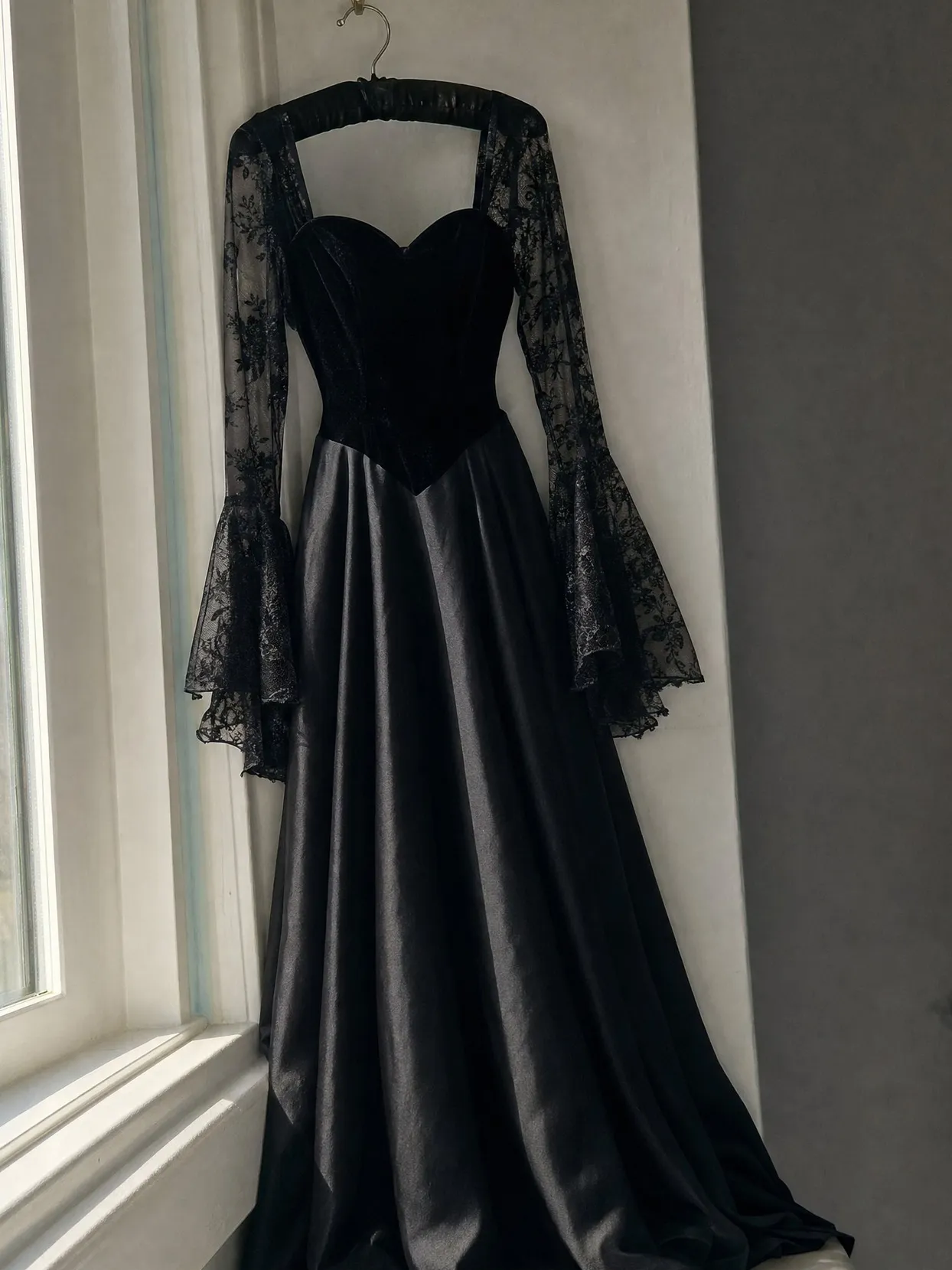 A black velvet bodice gown with sheer black lace sleeves and a satin skirt on a hanger by a window.