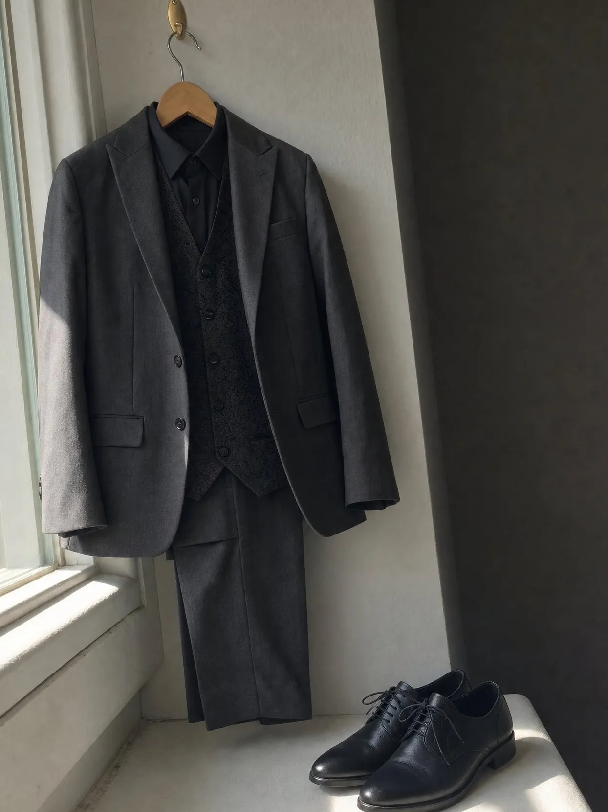 A charcoal suit with a black brocade waistcoat and a black shirt on a hanger by a window, shoes on the sill.