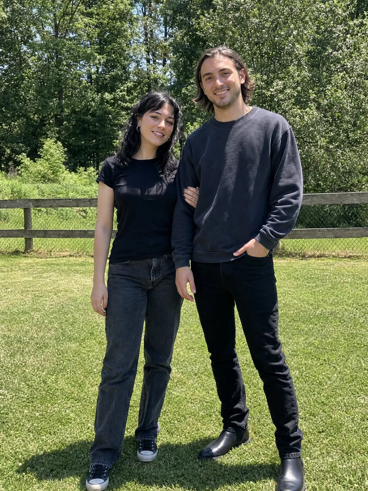 A couple standing arm in arm on a lawn by a wooden fence, in black and gray everyday clothes.