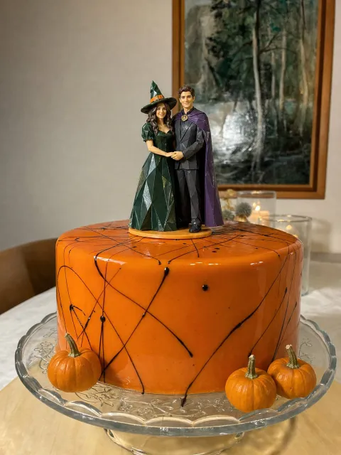 Isang couple topper, siya ay naka-berdeng witch hat at gown, siya naman ay naka-gray na suit at purple na kapa, sa orange na cake na may maliliit na kalabasa.