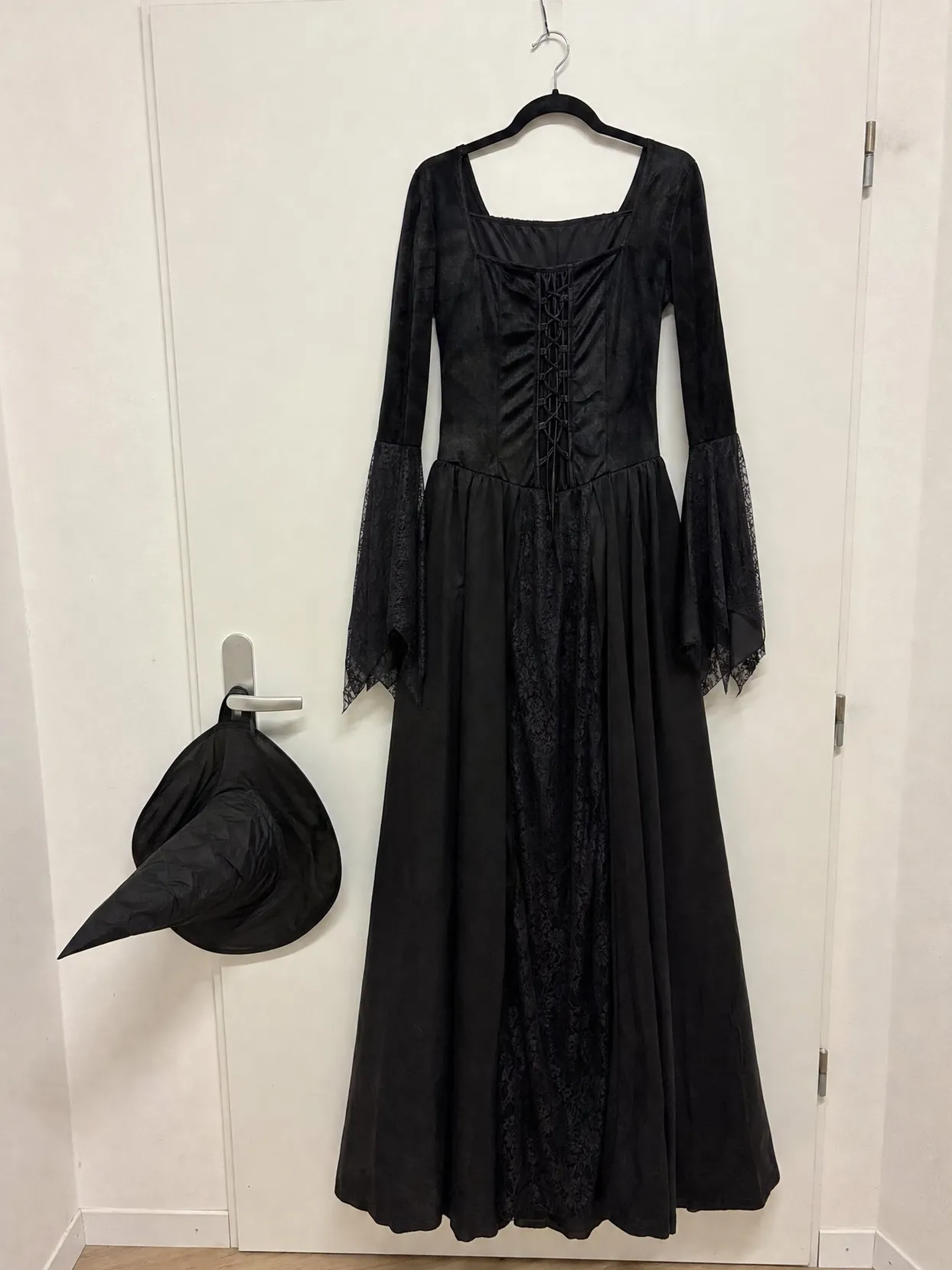 A long black dress with lace sleeves hanging on a white door, a black witch hat on the handle.