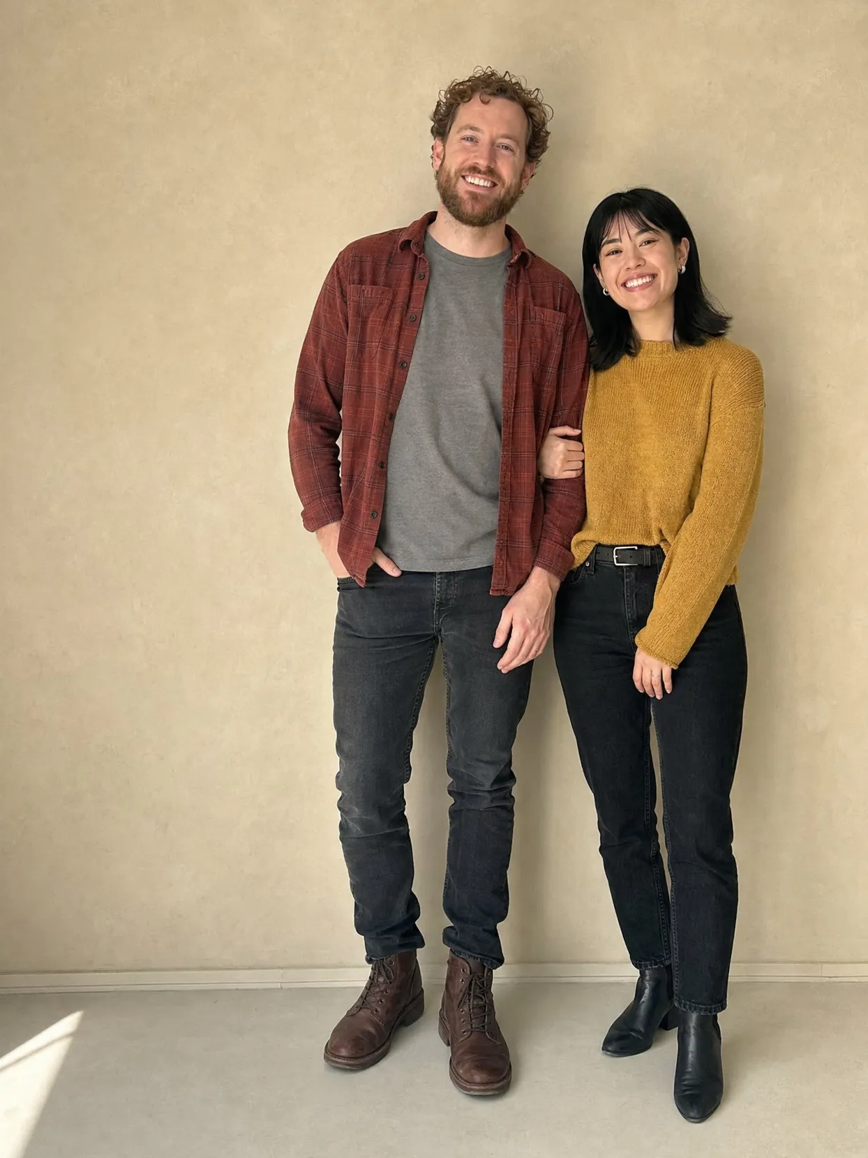A couple standing against a beige wall, he in a plaid shirt and jeans, she in a mustard sweater and black jeans.