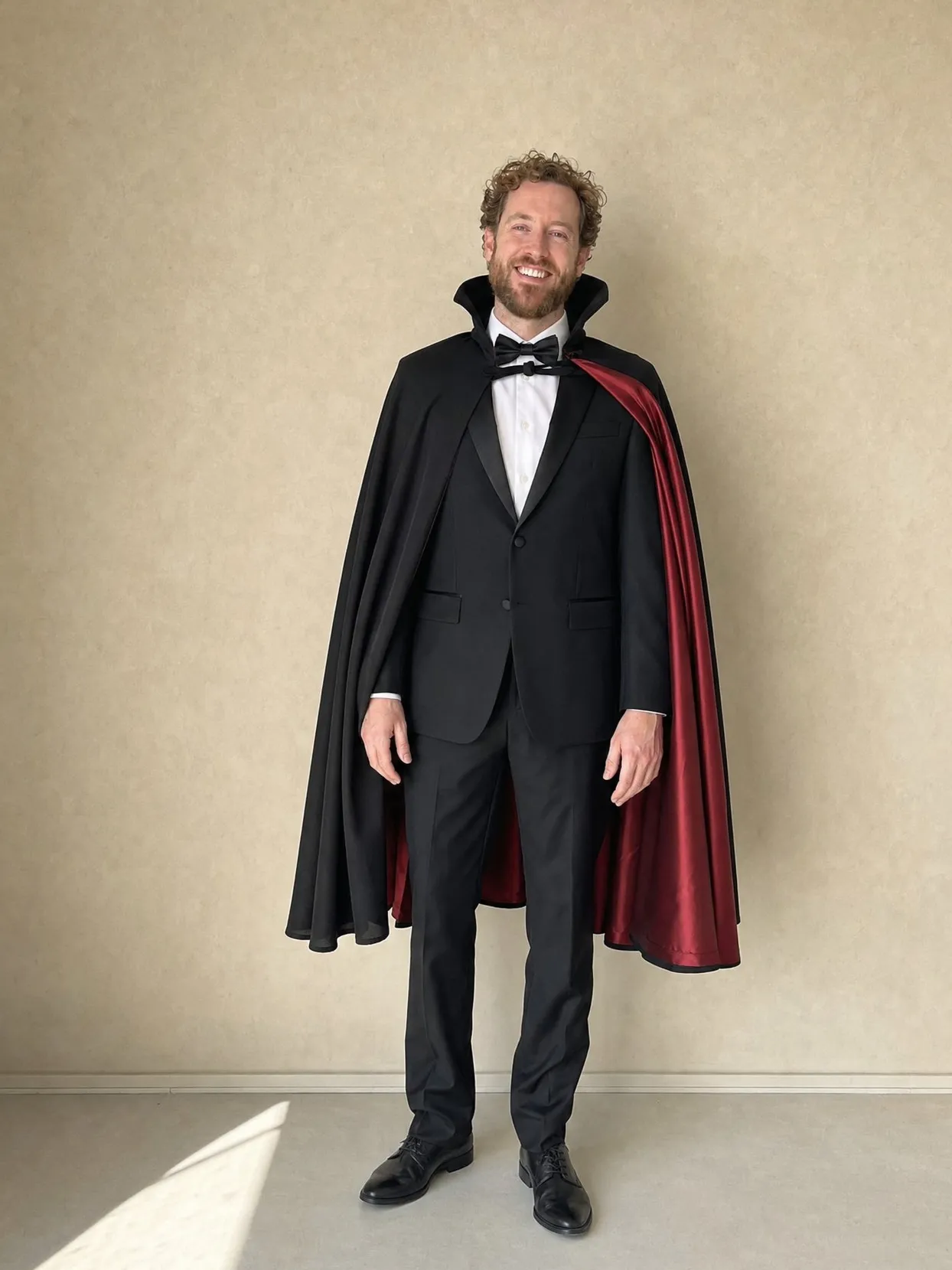 A man in a tuxedo and a black cape with a red lining standing against a beige wall.