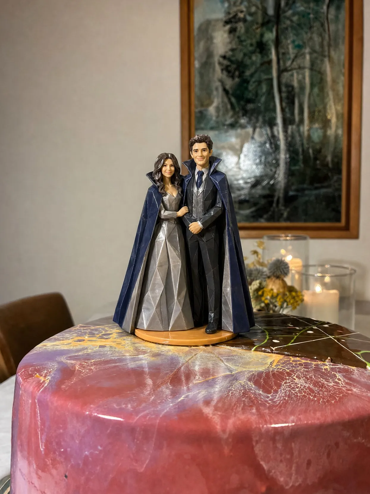 A couple topper, both in high-collar blue capes, she in a silver gown, he in a charcoal suit, on a pink and burgundy cake.