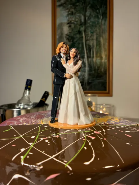 Printed bride and groom figure on an orange round base on top of a chocolate glazed cake, candles behind