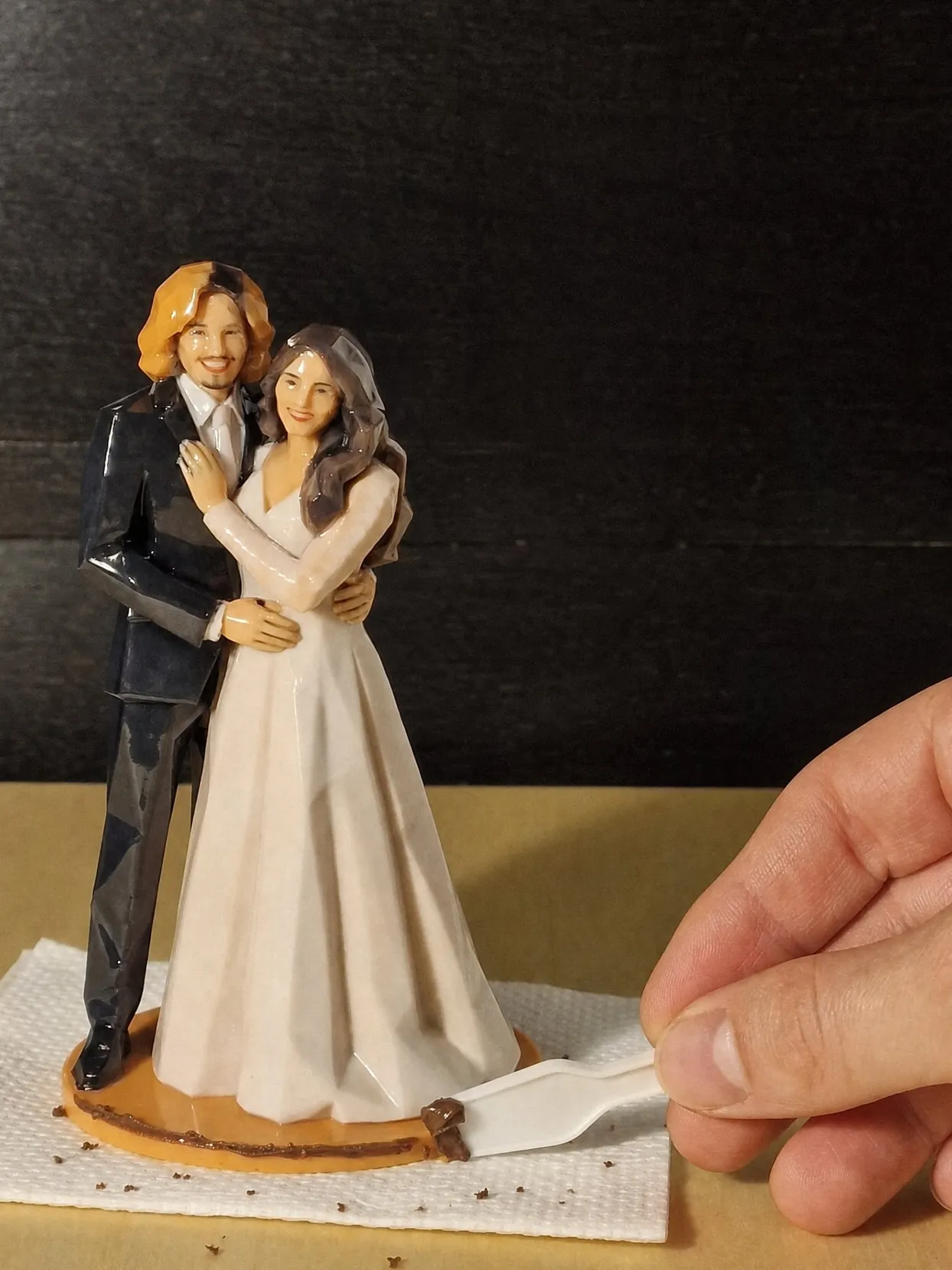 Bride and groom figure on a paper towel while a hand scrapes chocolate frosting off its orange base with a white plastic spatula
