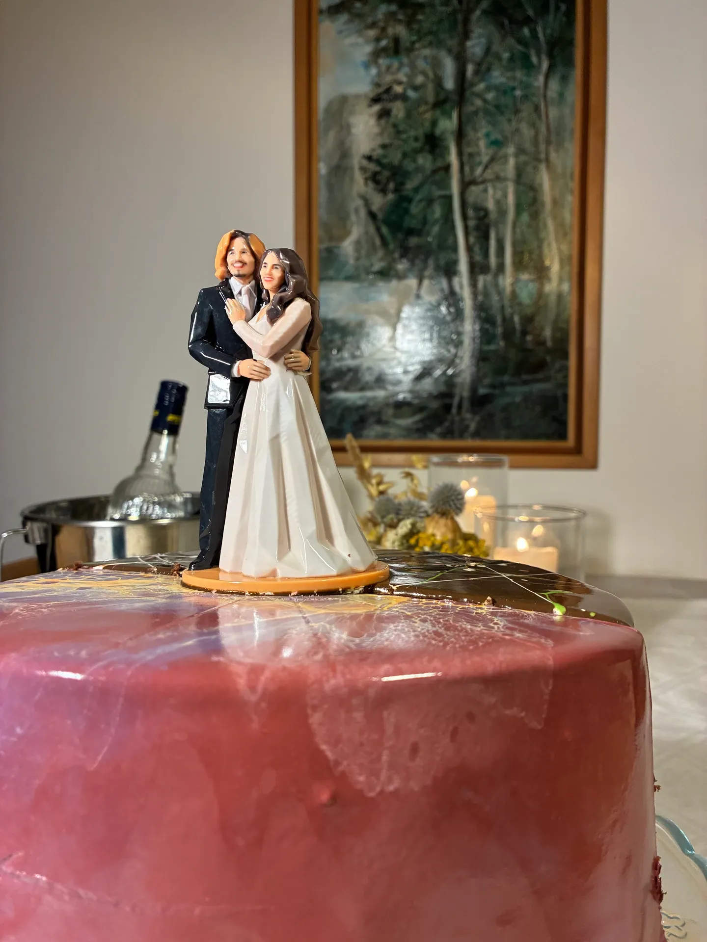Printed bride and groom figure on a round orange base on top of a pink glazed cake, seen at the height of the figure