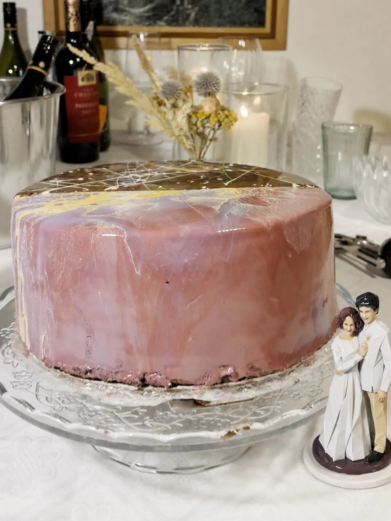 Bride and groom figure on a small white round board on a tablecloth beside a pink glazed cake