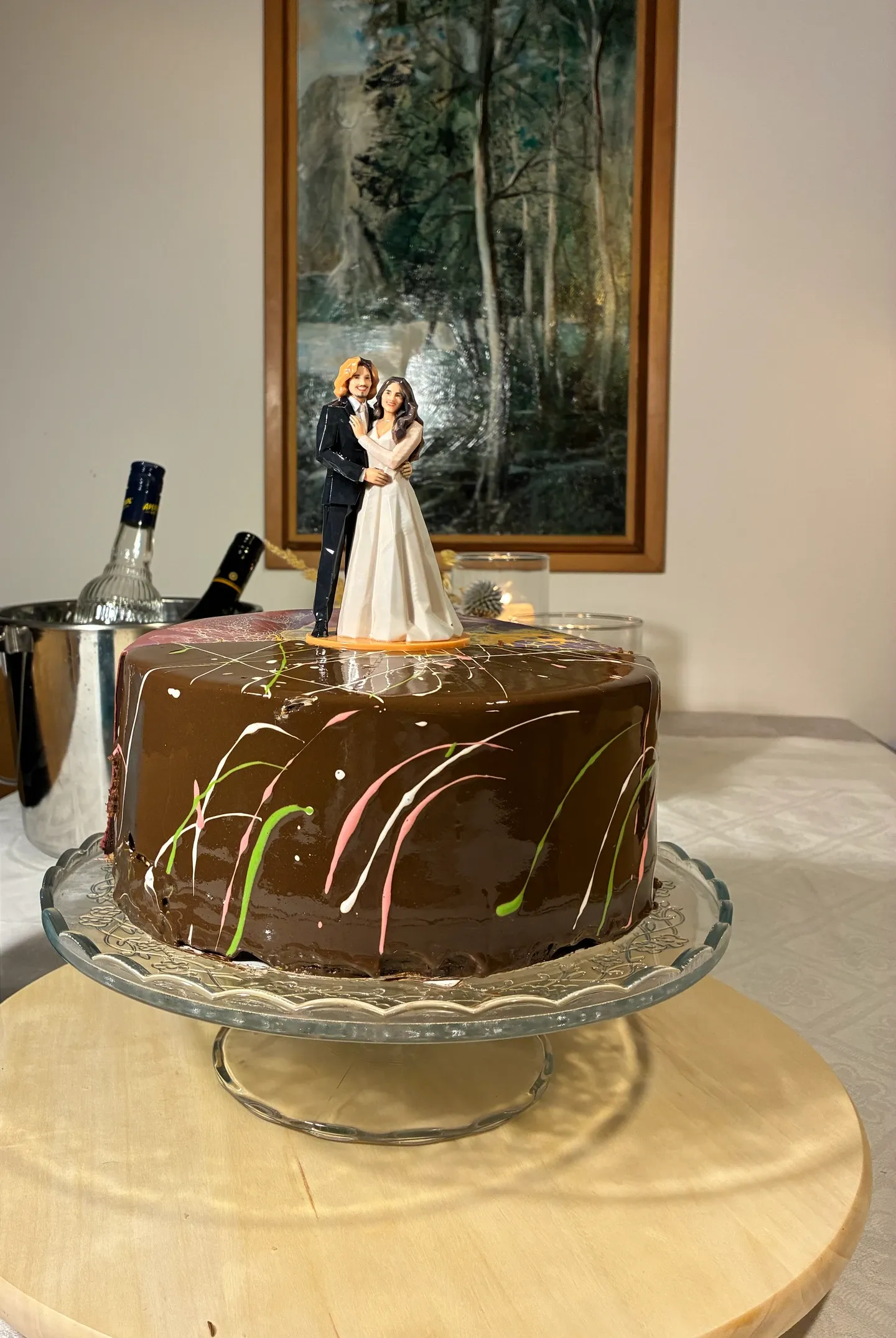 Chocolate cake on a glass stand with a small couple figure on top, an ice bucket with bottles and a large painting behind it