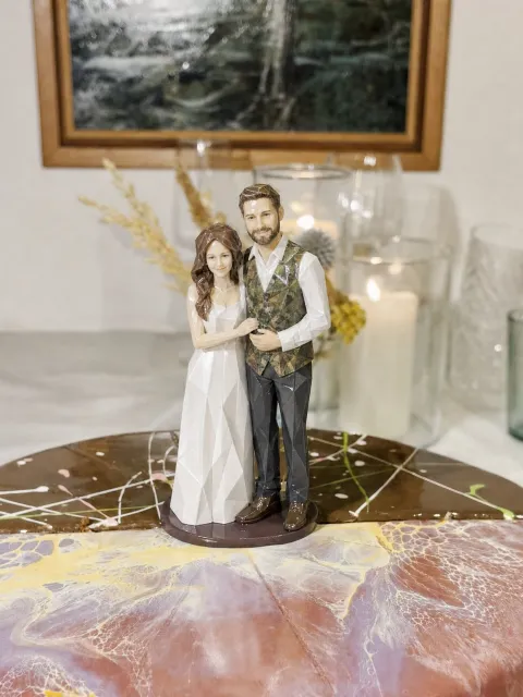 Origami na topper ng isang groom na naka-berdeng camo vest sa ibabaw ng puting polo at gray na pantalon, at isang bride na naka-puting gown, sa ibabaw ng marbled cake.