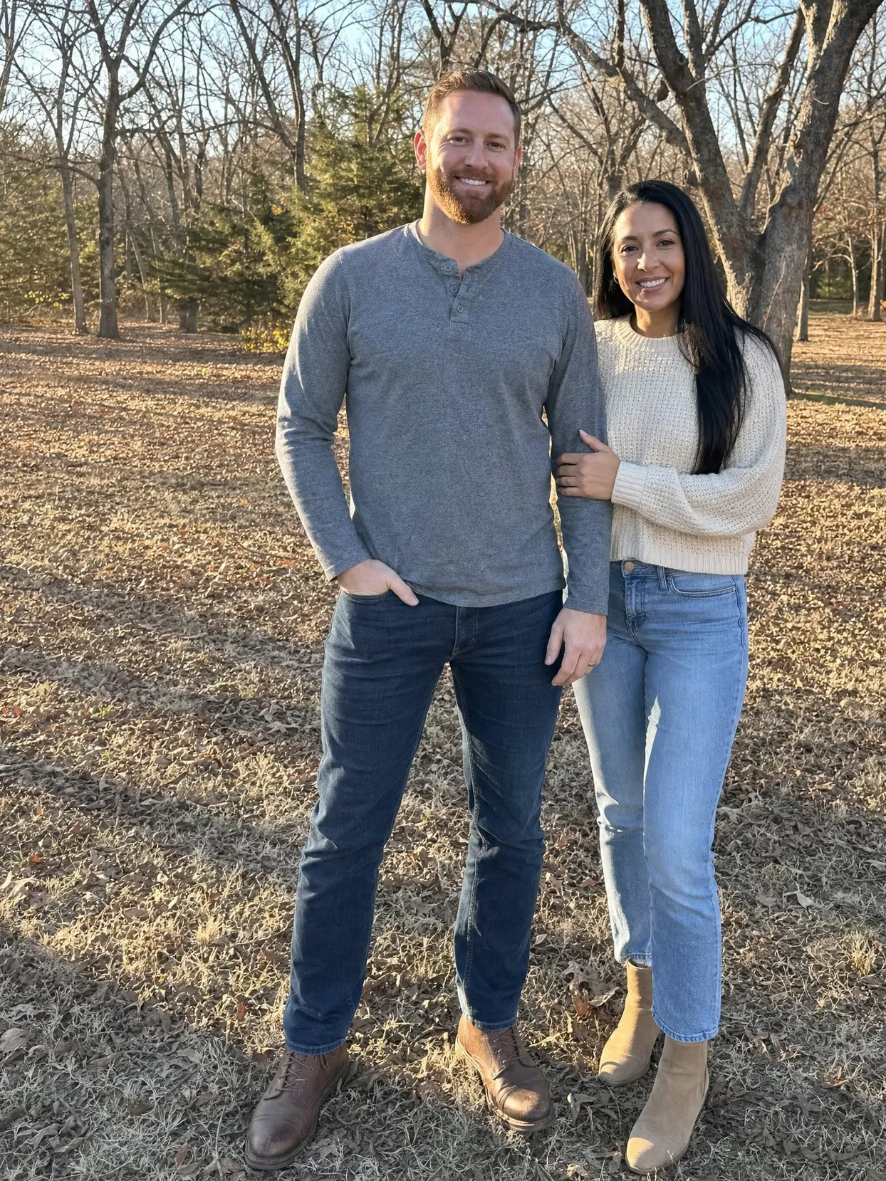 Ein Paar in einem kahlen Herbstwäldchen bei tiefer Sonne, er in grauem Henley-Shirt und Jeans, sie in cremefarbenem Pullover.
