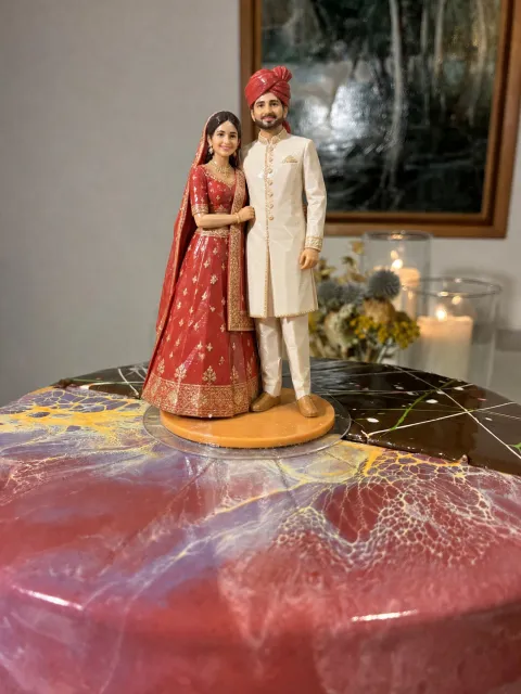 Topper ng Indian bride na naka-pulang lehenga at groom na naka-ivory na sherwani at pulang turban, sa pink at purple na glazed cake.