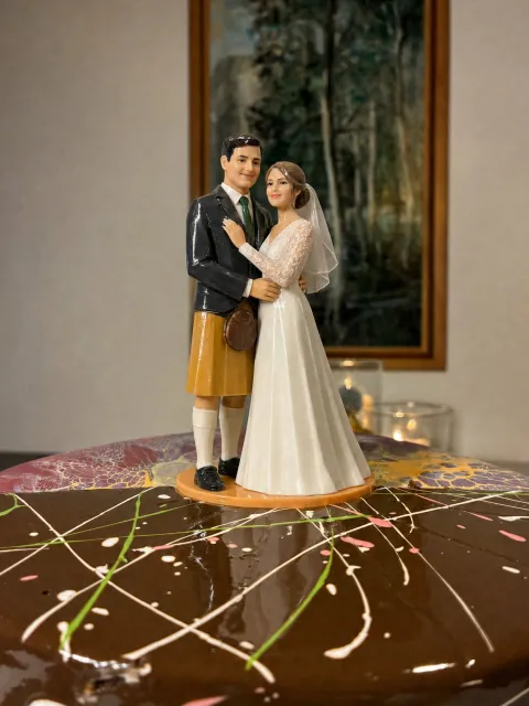 Topper ng groom na naka-saffron na kilt, madilim na jacket at green na kurbata at bride na naka-white gown na may lace na manggas at belo, sa chocolate cake na may pink, puti at green na drizzle.