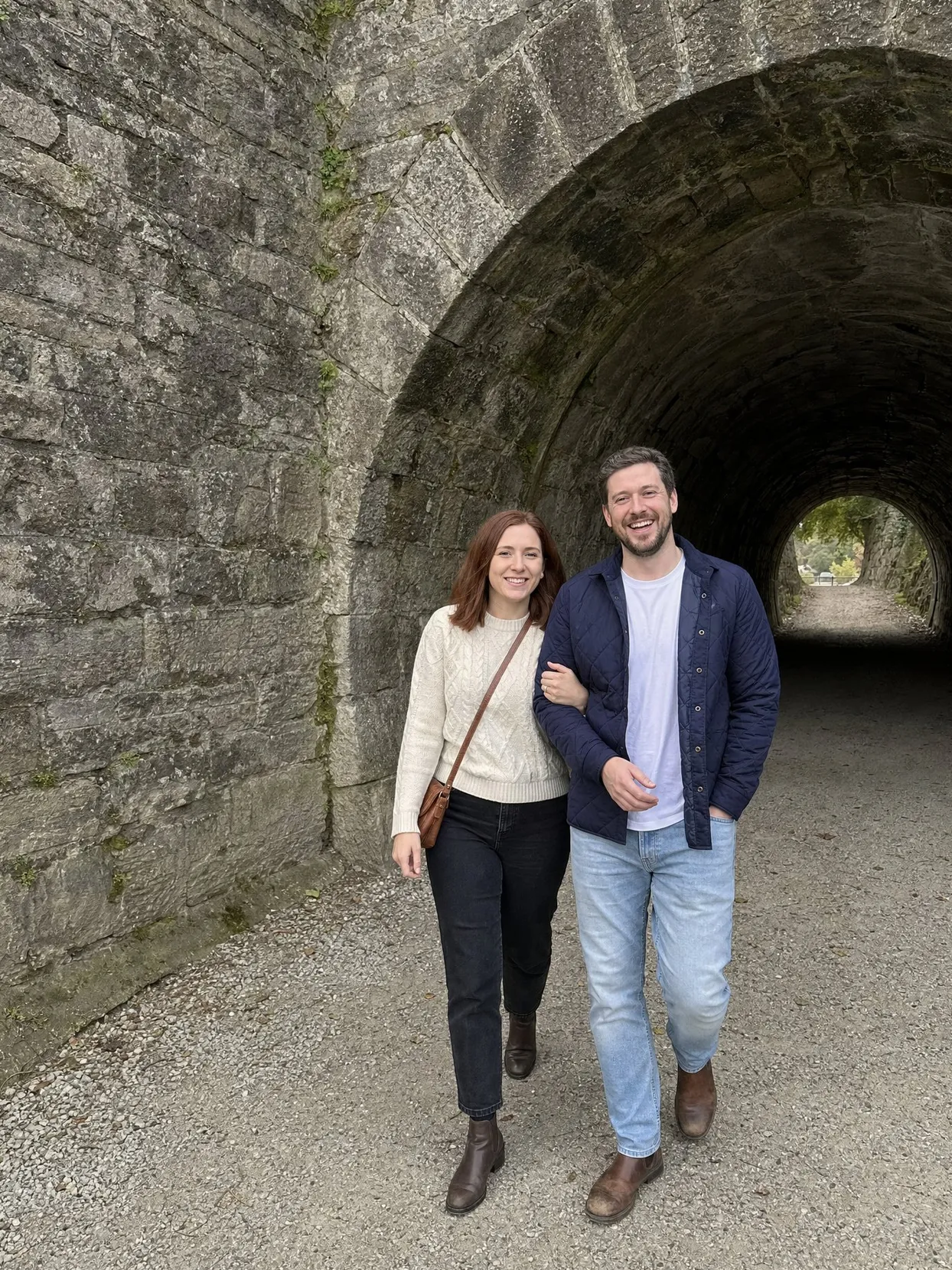 A couple walking arm in arm out of an old stone tunnel on a gravel path, he in a navy quilted jacket and jeans, she in a cream cable-knit sweater and black jeans.