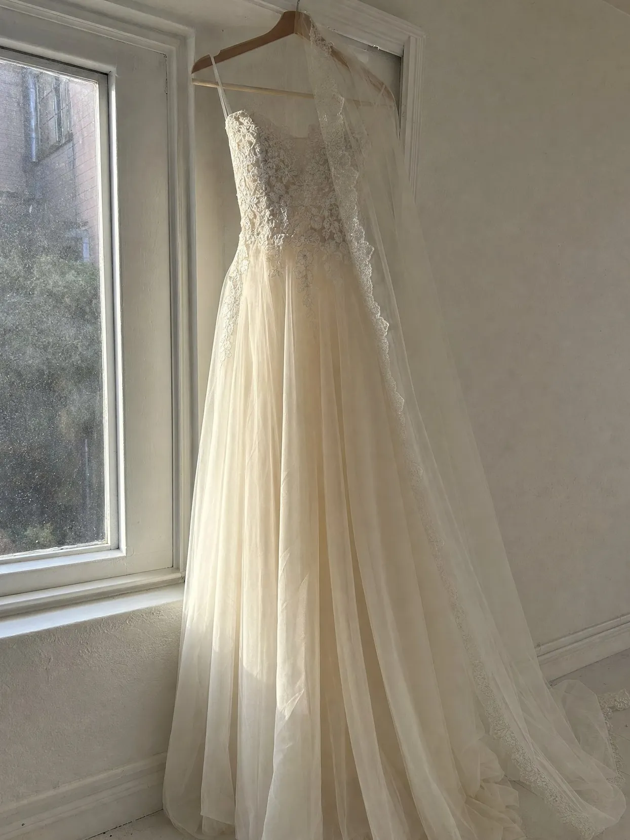 A sleeveless ivory lace wedding gown and a long veil with a lace edge on a wooden hanger by a sunny window.