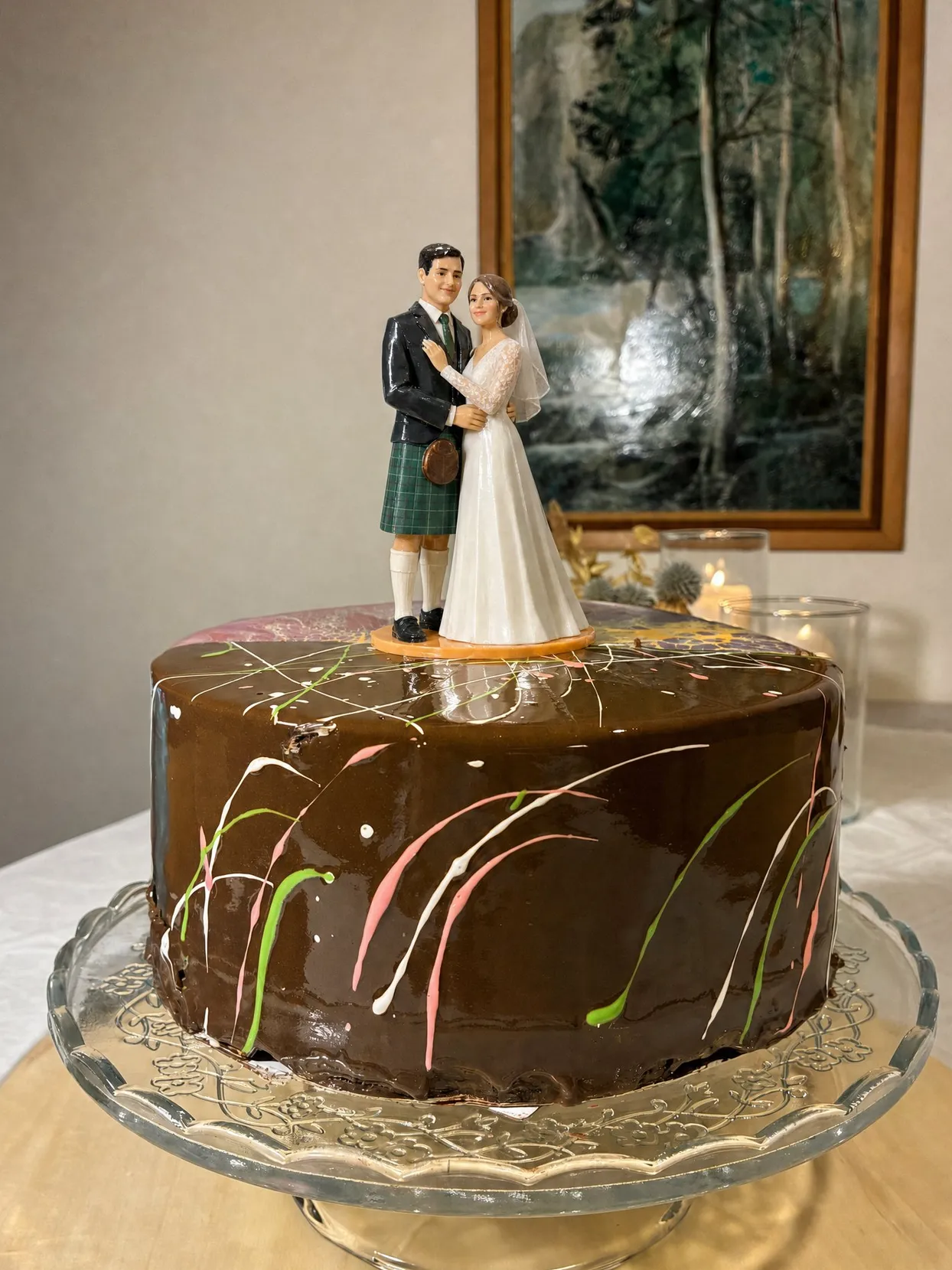 The same topper couple, the groom in a green tartan kilt, on a whole chocolate drip cake on a glass cake stand.