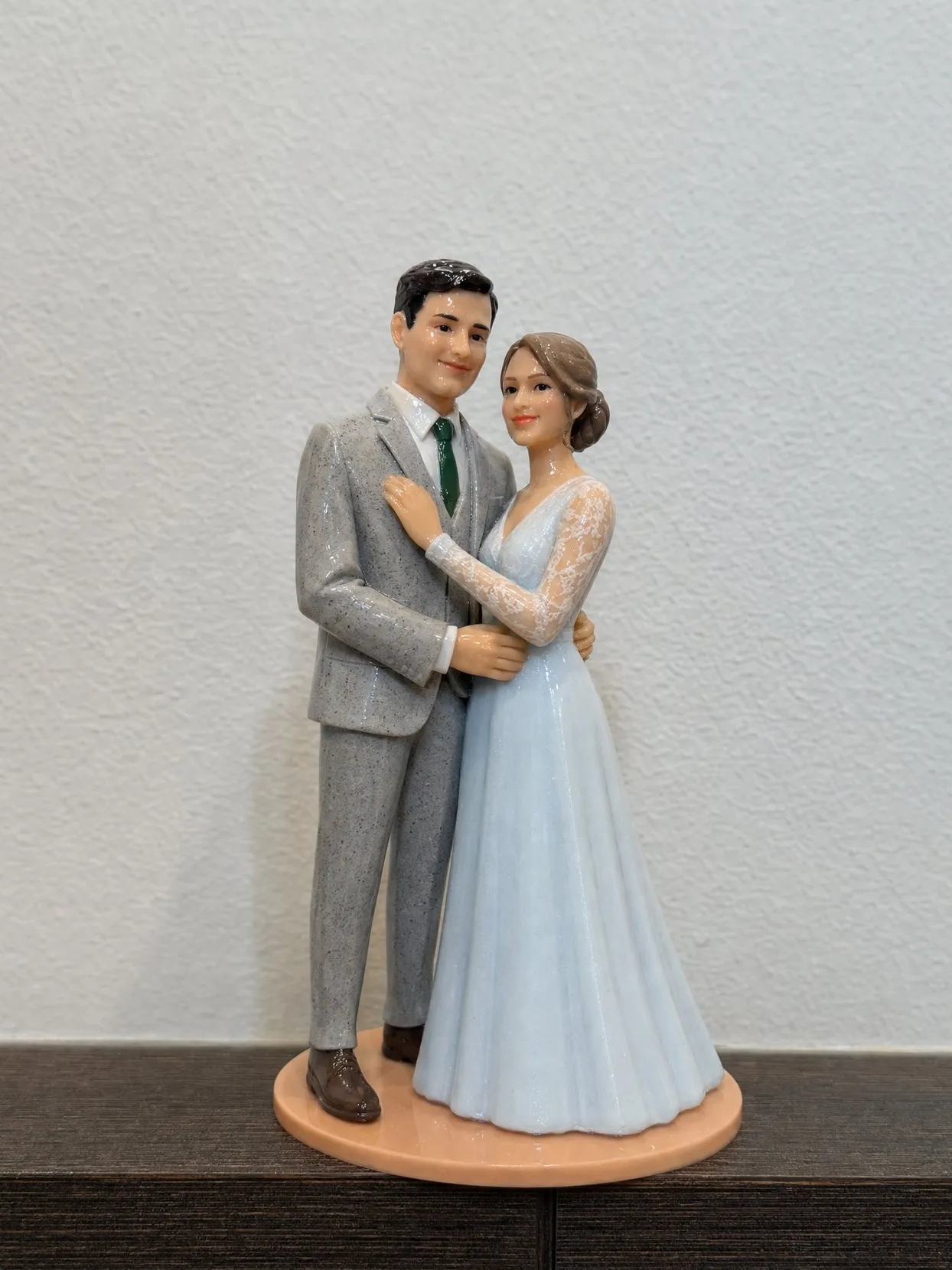 The same topper couple, the groom in a gray tweed suit, the bride in a pale blue gown with lace sleeves, on a dark wooden sideboard against a white wall.